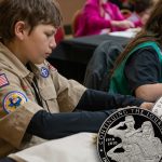 A Boy Scout and a Girl Scout studying at a table with the U.S. Mint Boy Scout and Girl Scout commemorative coins.